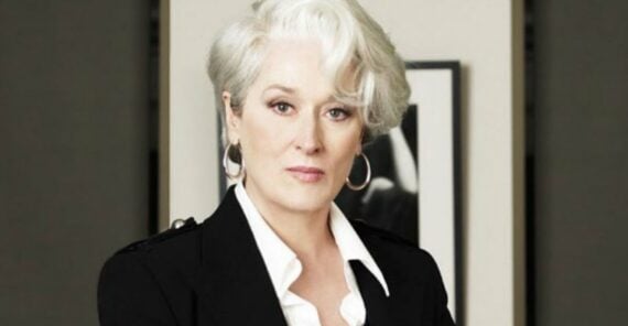 A woman with short, stylish white hair and hoop earrings wears a black blazer over a white shirt, looking confidently at the camera. The background is blurred, showing a black-and-white photo on the wall.