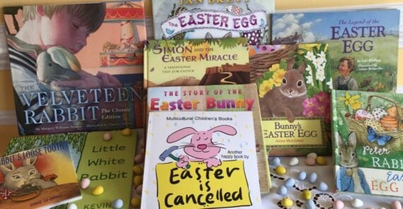 A collection of colorful children's Easter books, including titles like "The Velveteen Rabbit" and "Easter is Cancelled," is spread out on a table with pastel candy eggs scattered in front.