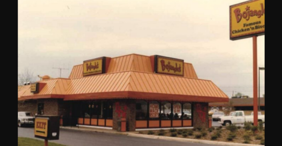 A Bojangles' restaurant with a brown and orange roof sits beside a parking lot with several cars. A large sign displays the Bojangles' logo and the words "Famous Chicken 'n Biscuits.