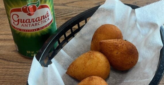 A black basket lined with white paper holds four golden-brown coxinhas. Next to the basket is a can of Guaran&aacute; Antarctica soda on a wooden table.