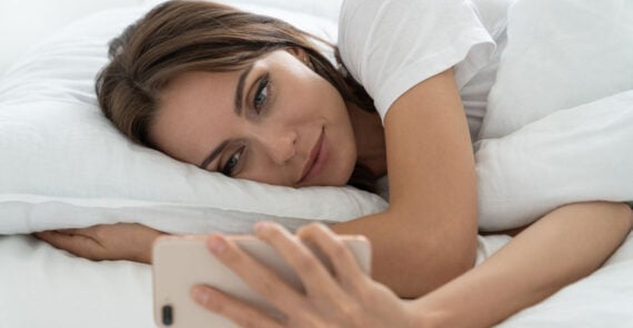 A woman lies in bed under white sheets, resting her head on a pillow while smiling and looking at a smartphone she is holding in her hand.