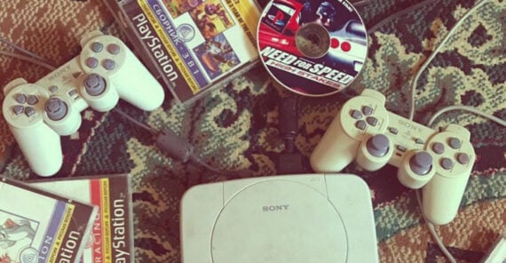 A white Sony PlayStation console with two controllers, PlayStation game cases, and a Need for Speed game disc are on a patterned carpet.