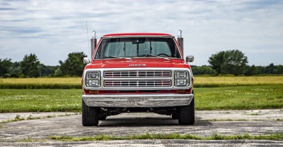 A red vintage Dodge pickup truck is parked on a paved area, facing the camera, with a grassy field and trees visible in the background under a partly cloudy sky.