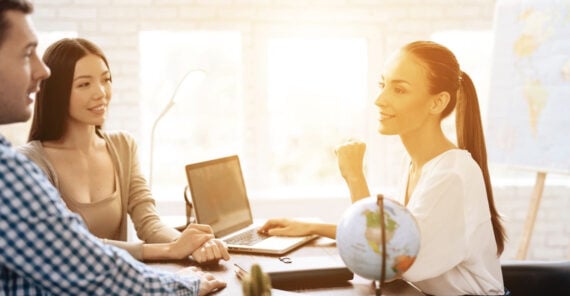 Three people sit at a table in a bright office, having a discussion. A woman with a laptop faces a man and another woman. A small globe and office supplies are on the table, and sunlight streams in from large windows.