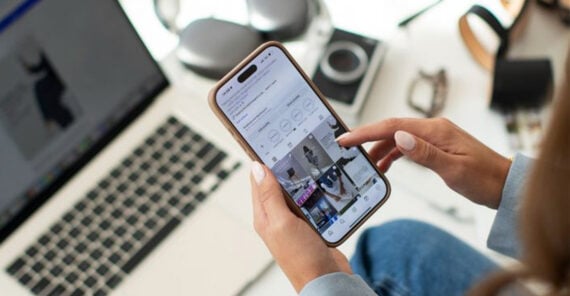 A person holds a smartphone and scrolls through a social media profile, with a laptop, headphones, and office supplies visible on a white desk in the background.