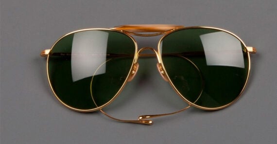 A pair of gold-framed aviator sunglasses with dark green lenses is displayed on a grey background. The sunglasses have a double bridge and thin metal arms.