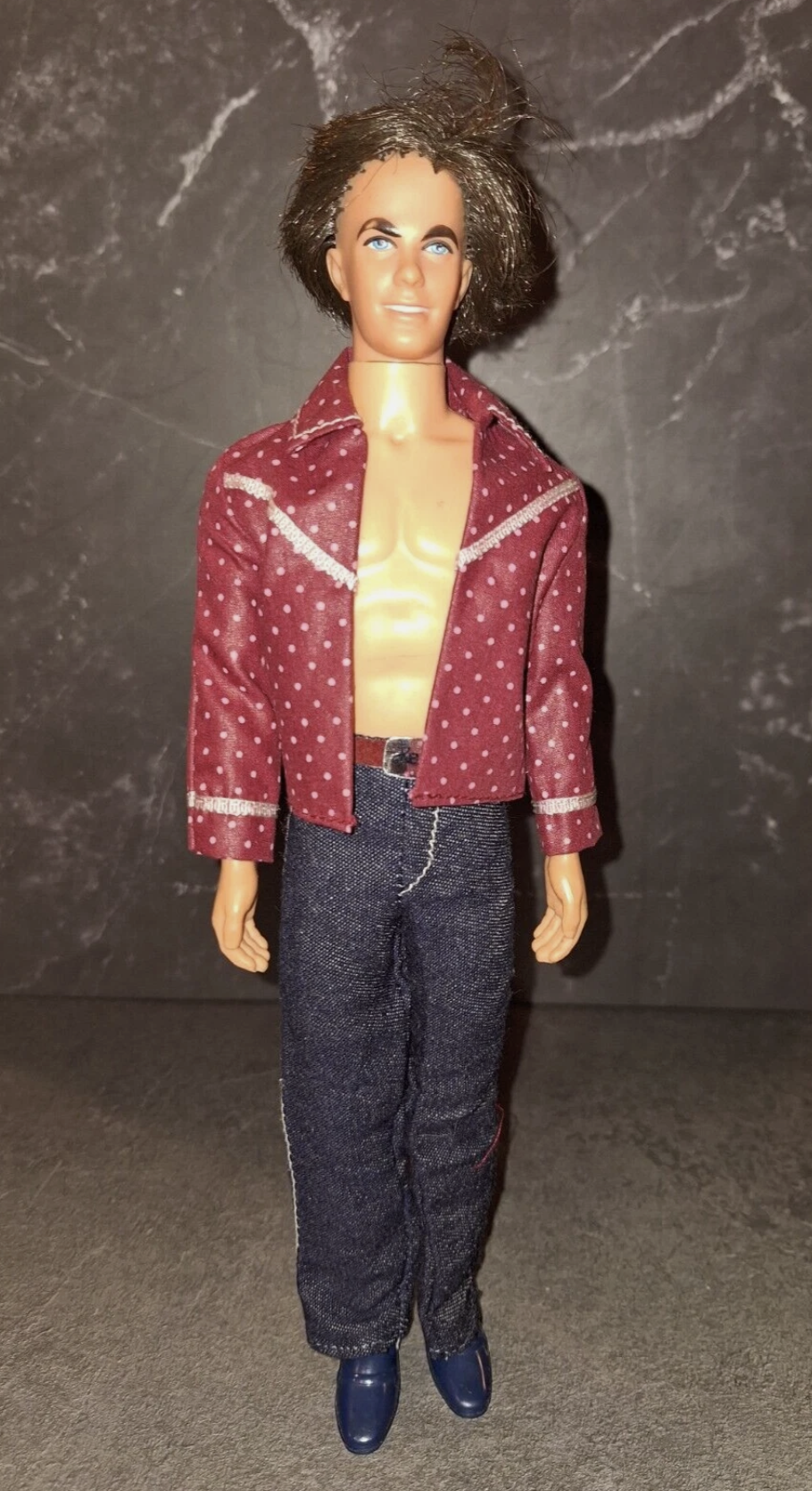 A vintage male doll with blue eyes, tousled brown hair, a maroon polka-dotted jacket, no shirt, dark blue jeans, and blue shoes stands against a gray textured background.