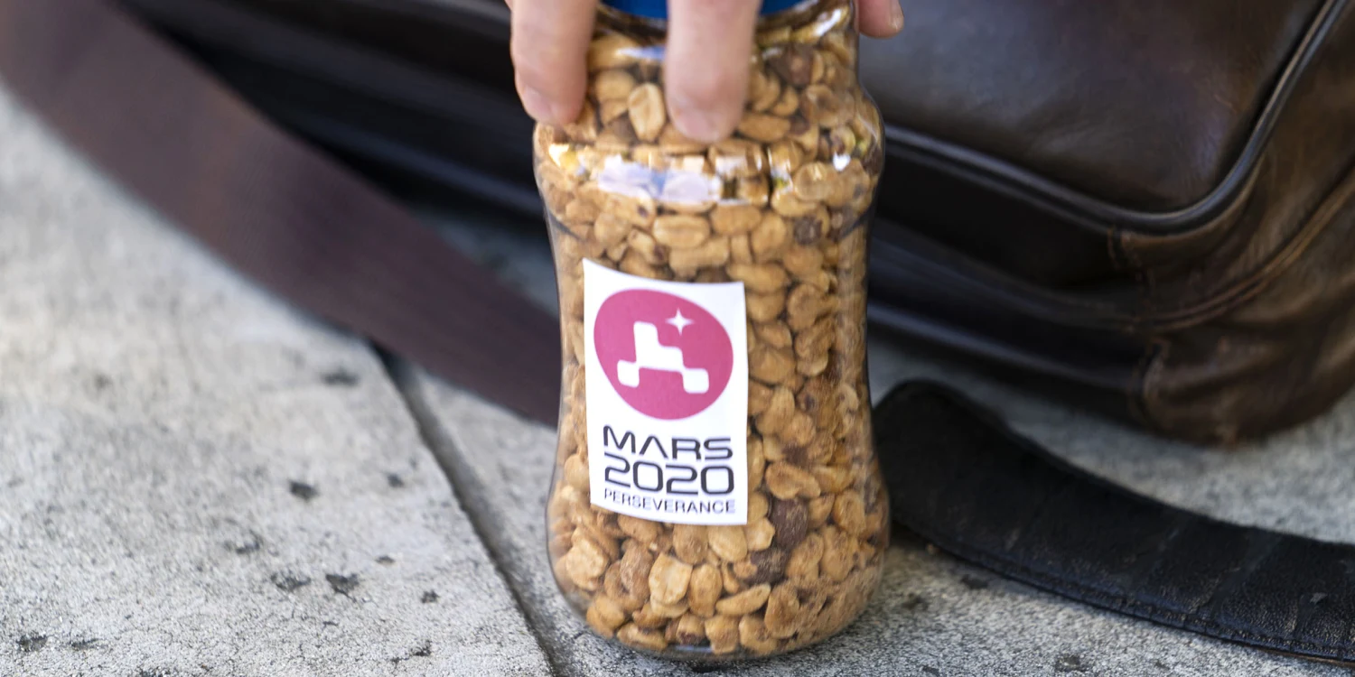 A hand holds a jar filled with peanuts, labeled with a Mars 2020 Perseverance mission sticker, resting on a sidewalk next to a brown bag.