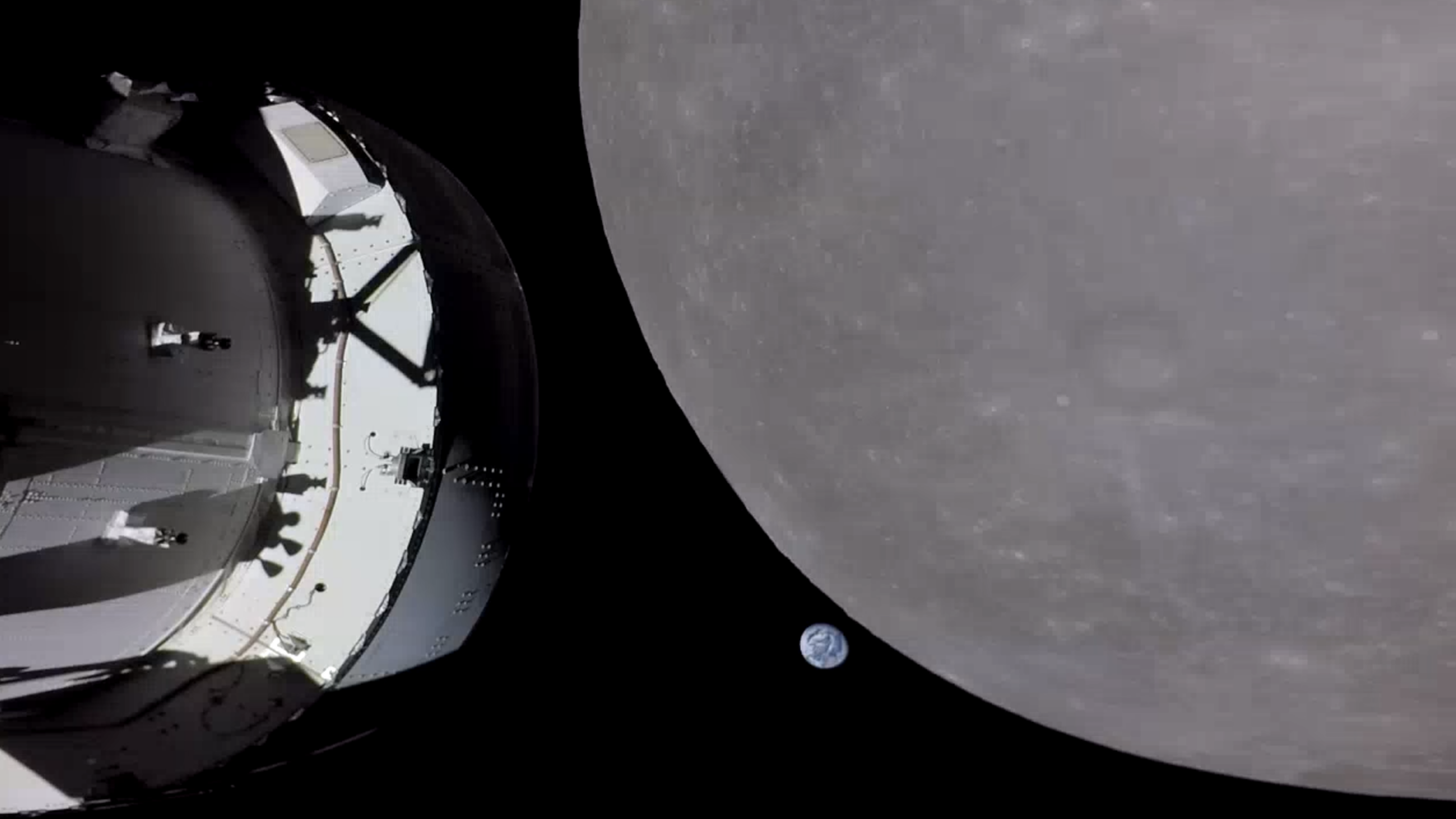 A spacecraft orbits the Moon, with part of the spacecraft visible on the left. The large, gray surface of the Moon dominates the right side, and a small, distant Earth appears against the blackness of space.