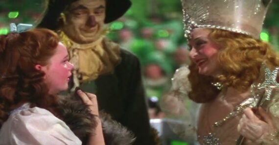 Dorothy, holding Toto, talks to Glinda the Good Witch, who wears a sparkling crown and holds a wand, while the Scarecrow stands behind them. All are characters from "The Wizard of Oz.