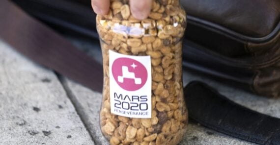 A hand holds a clear plastic jar filled with peanuts. The jar has a "Mars 2020 Perseverance" sticker showing the rover&rsquo;s logo. The jar rests on a concrete surface next to a brown leather bag.