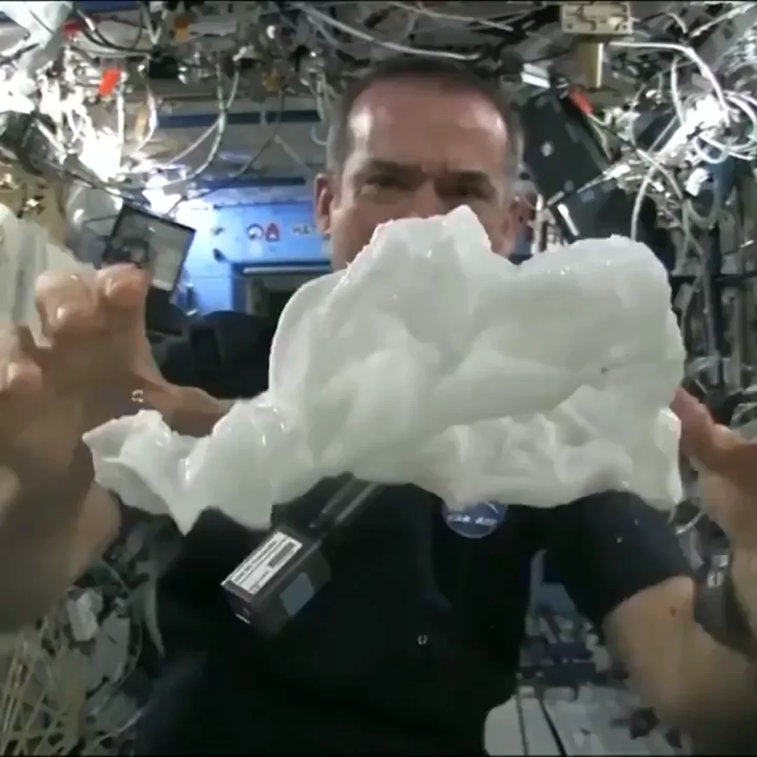 An astronaut in a space station floats a large blob of water in mid-air, surrounded by equipment, wires, and machinery in a microgravity environment.