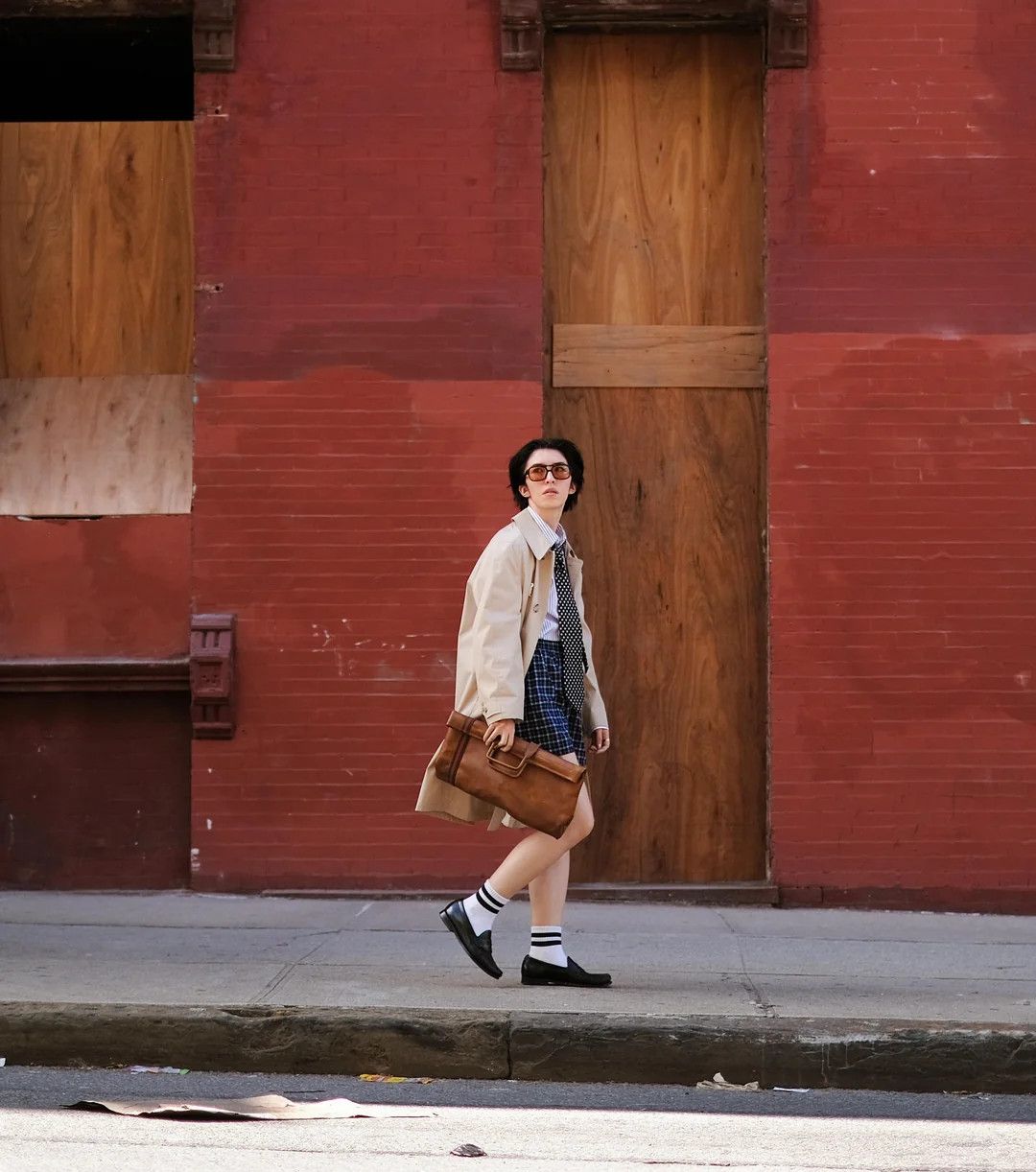 A person wearing sunglasses, a beige coat, and a blue dress walks on a sidewalk beside a red brick building with boarded-up windows and doors, carrying a brown bag.