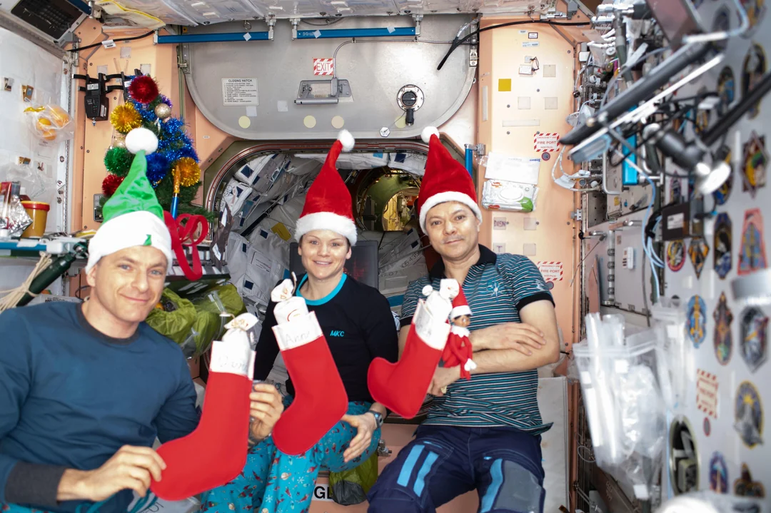 Three astronauts in festive hats sit inside a space station decorated for Christmas, smiling and holding red stockings. The background features equipment, colorful ornaments, and mission patches on the wall.