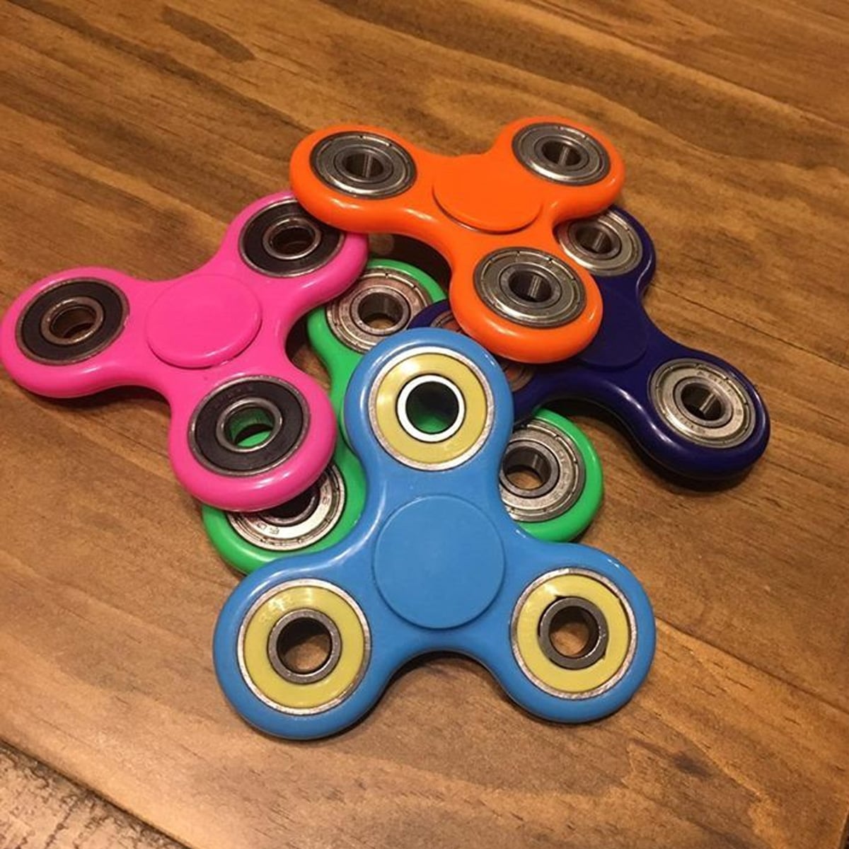 Five colorful fidget spinners—pink, green, blue, orange, and navy—are stacked on a wooden surface, with their metal bearings visible in the center and on each arm.