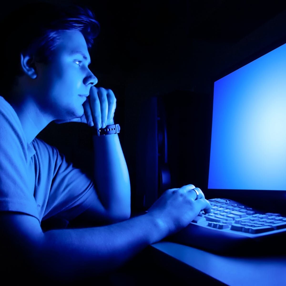 A person sits in a dark room, illuminated by a blue glow from a computer monitor, looking intently at the screen with one hand on the keyboard and the other resting on their chin.