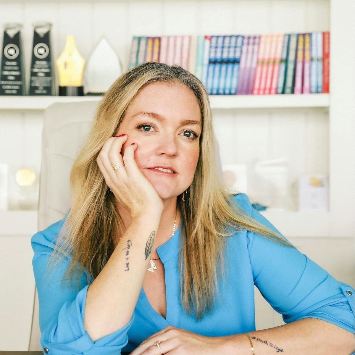 A woman with long blonde hair, wearing a light blue blouse, rests her head on her hand at a desk. Books and awards are displayed on a white shelf in the background. She has tattoos on her arm and a relaxed expression.