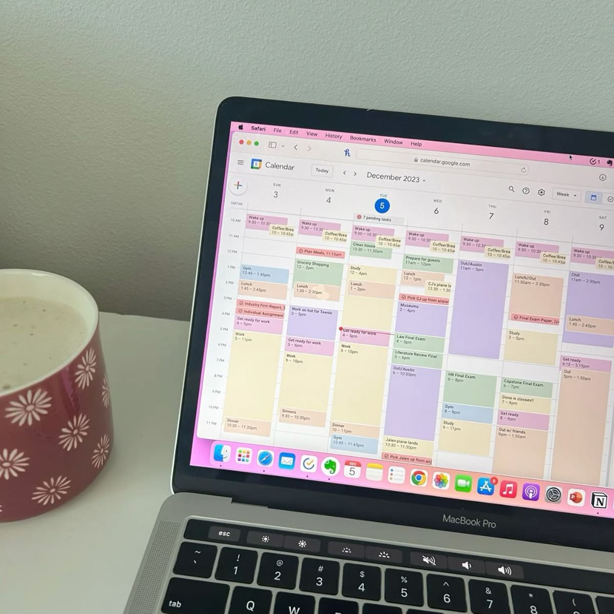 A laptop displaying a color-coded digital calendar for December 2023 sits on a white desk next to a red mug with white floral patterns.