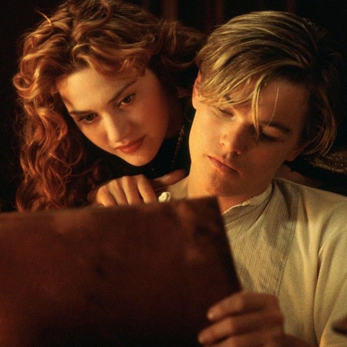 A young woman with curly red hair leans closely over a young man’s shoulder as they both look intently at a large brown sketchbook or notebook in soft, warm lighting.