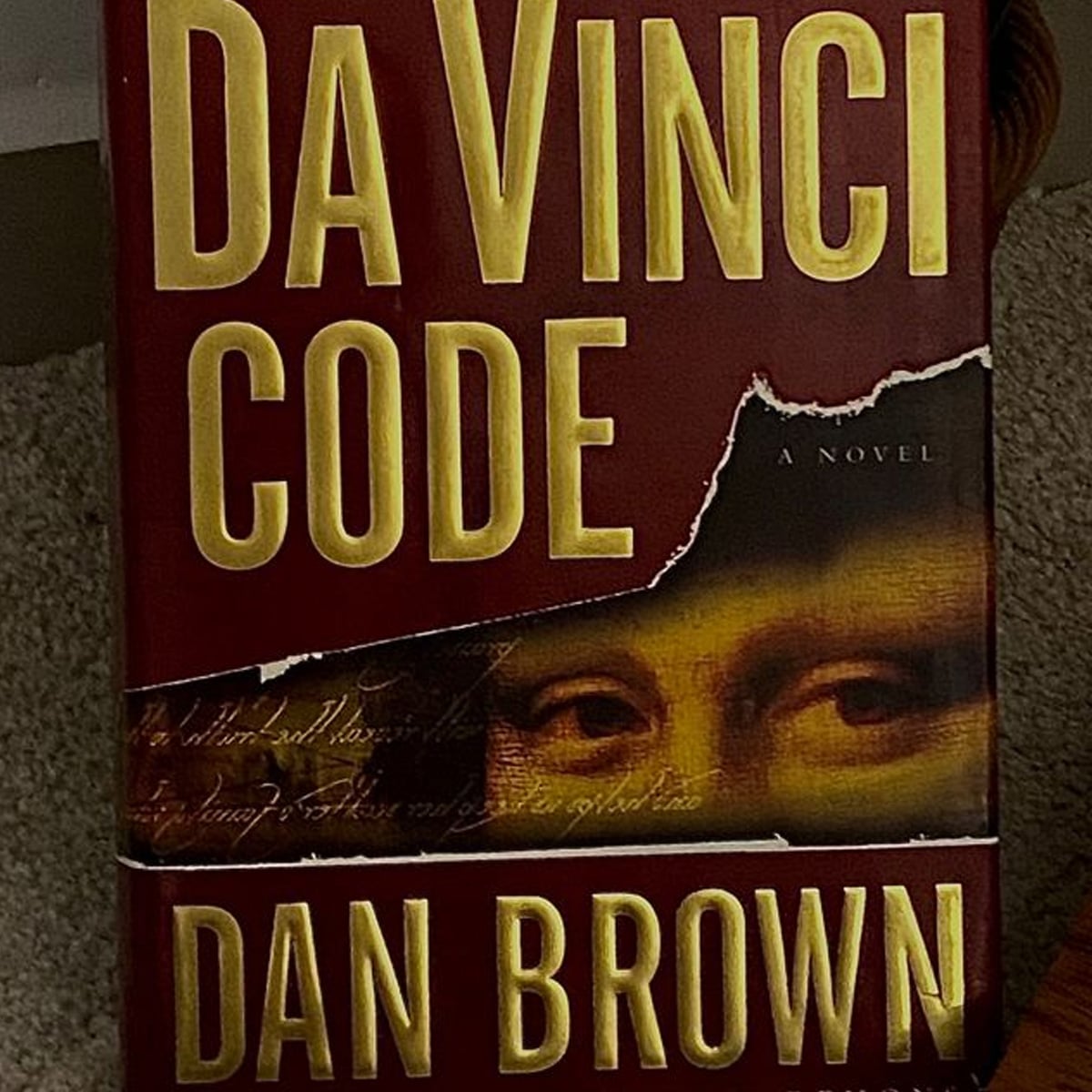 The cover of the book "The Da Vinci Code" by Dan Brown, featuring large yellow text over a red background with a torn effect that reveals part of the Mona Lisa’s face.