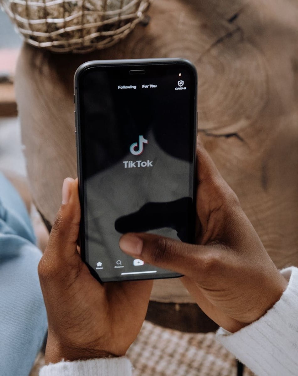 A person holds a smartphone displaying the TikTok app's home screen, with their thumb near the screen. The background shows part of a wooden table and another object out of focus.