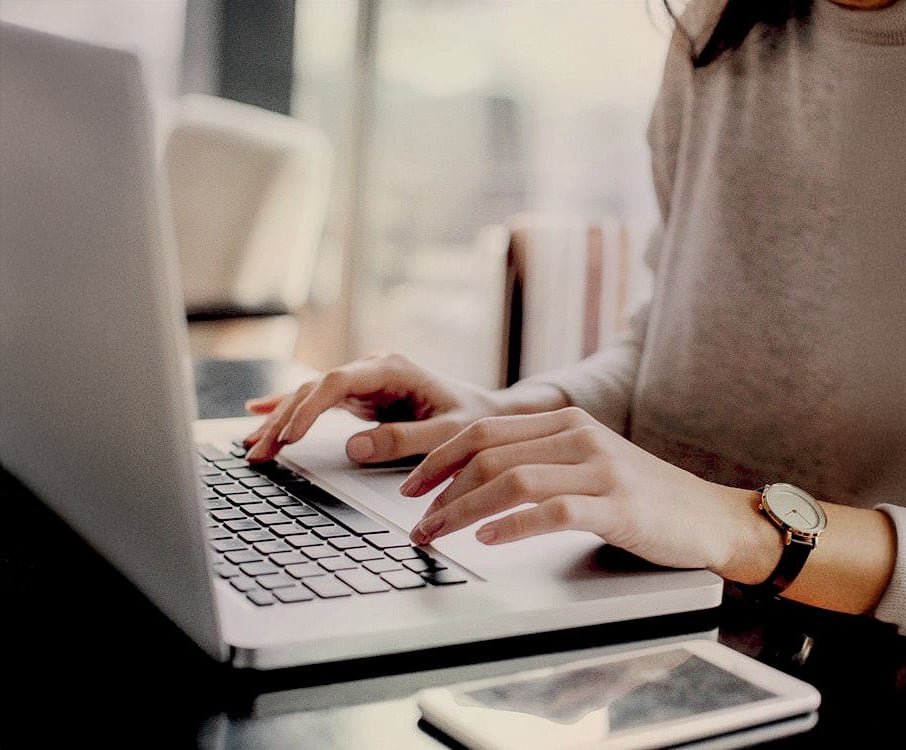 A person wearing a watch types on a laptop at a desk, with a smartphone placed nearby. The focus is on their hands and the keyboard, with a softly blurred background.