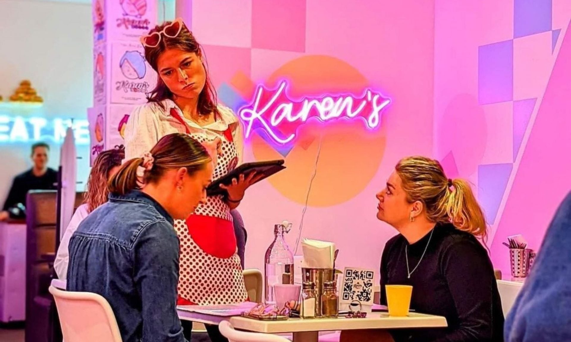 A waitress with a stern expression stands by a table where two women sit, one looking up at her. The cafe has bright pink decor and a neon sign reading “Karen’s” on the wall behind them.