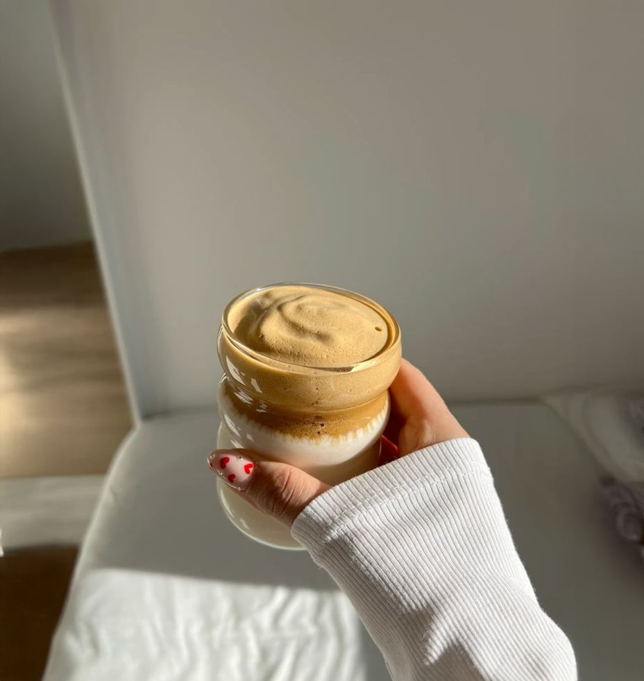 A hand with painted nails holds a glass of dalgona coffee, featuring whipped coffee on top of milk. The background is softly lit, showing a white bed and wall in a minimalistic room.