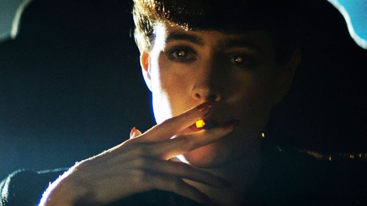 A person with short dark hair holds a lit cigarette near their lips, their face illuminated dramatically against a dark background, highlighting their intense gaze and red-painted nails.