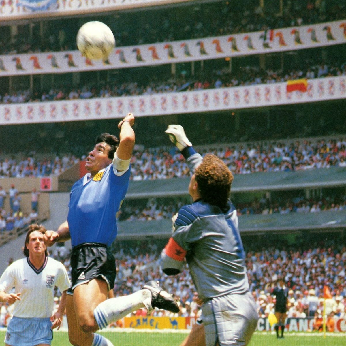 A soccer player in a blue jersey jumps and uses his hand to touch the ball near the goal as a goalkeeper in gray tries to reach it, with another player and a large crowd in the stadium background.