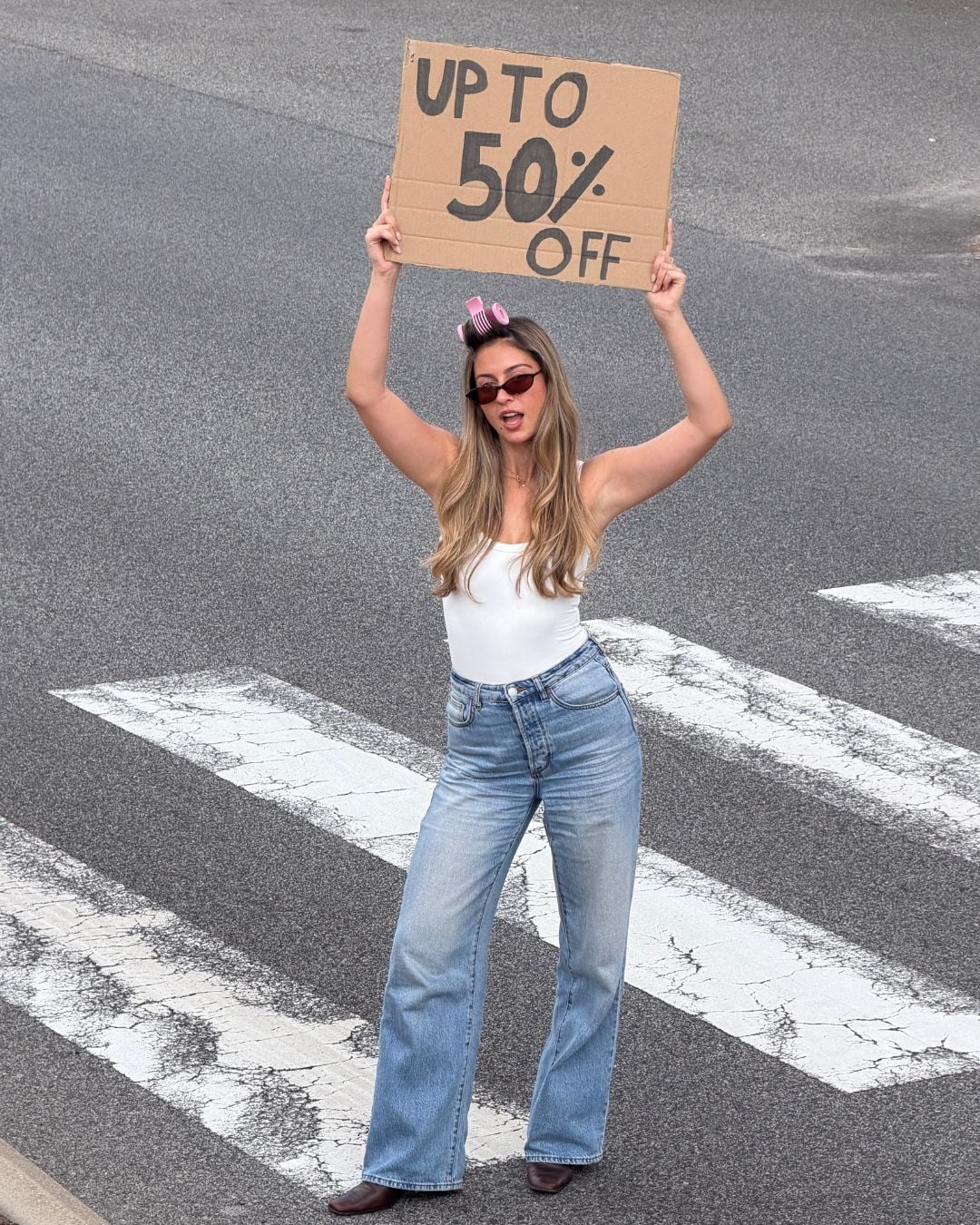 A woman wearing sunglasses, jeans, a white tank top, and hair rollers stands on a crosswalk holding a cardboard sign above her head that reads "UP TO 50% OFF.