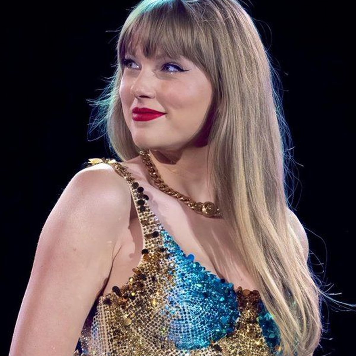 A woman with long blonde hair and red lipstick wears a gold and blue sequined dress, looking off to the side and smiling softly against a dark background. Artist.