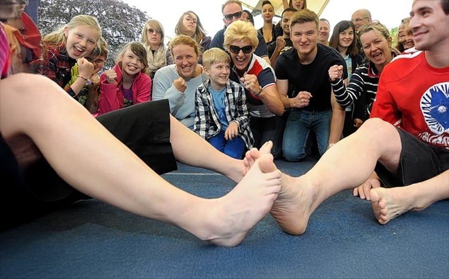 Two people compete in a toe wrestling match while a group of excited spectators, including children and adults, cheer and watch closely in the background.