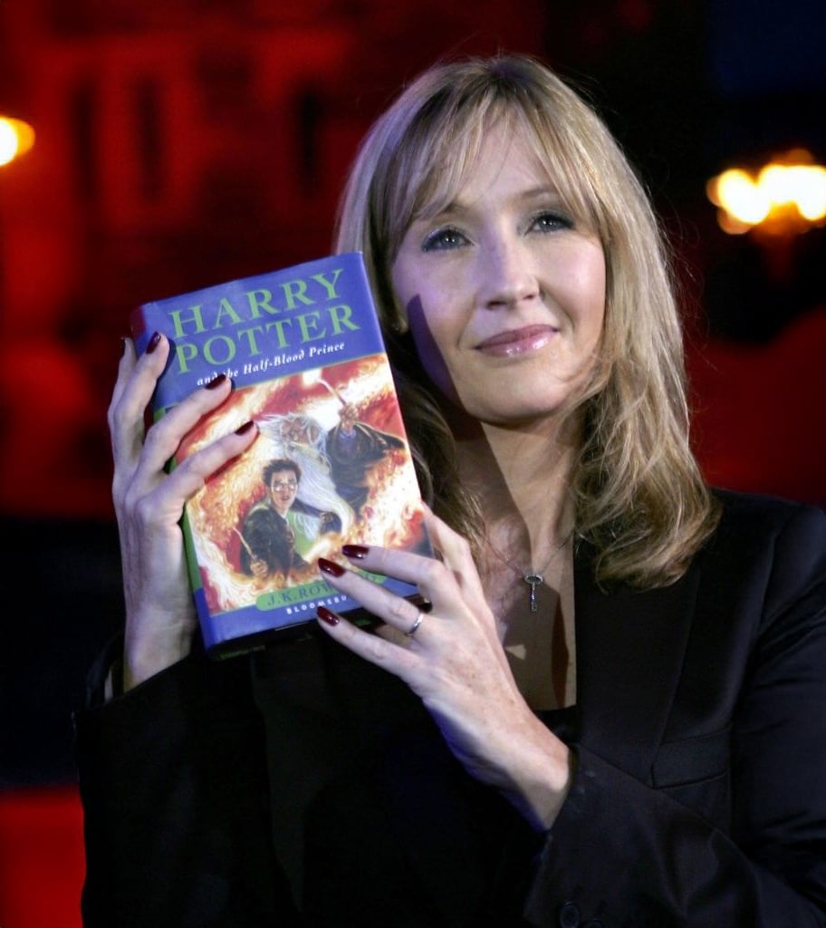 A woman with light brown hair wearing a black outfit holds up a copy of "Harry Potter and the Half-Blood Prince" and smiles, with a blurred red and blue background behind her.