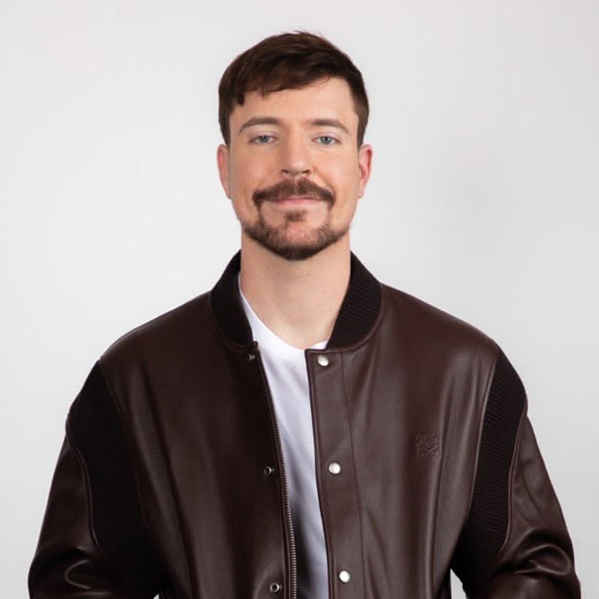 A man with short brown hair and a mustache is smiling slightly. He is wearing a dark brown leather jacket over a white shirt and is standing against a plain light background.