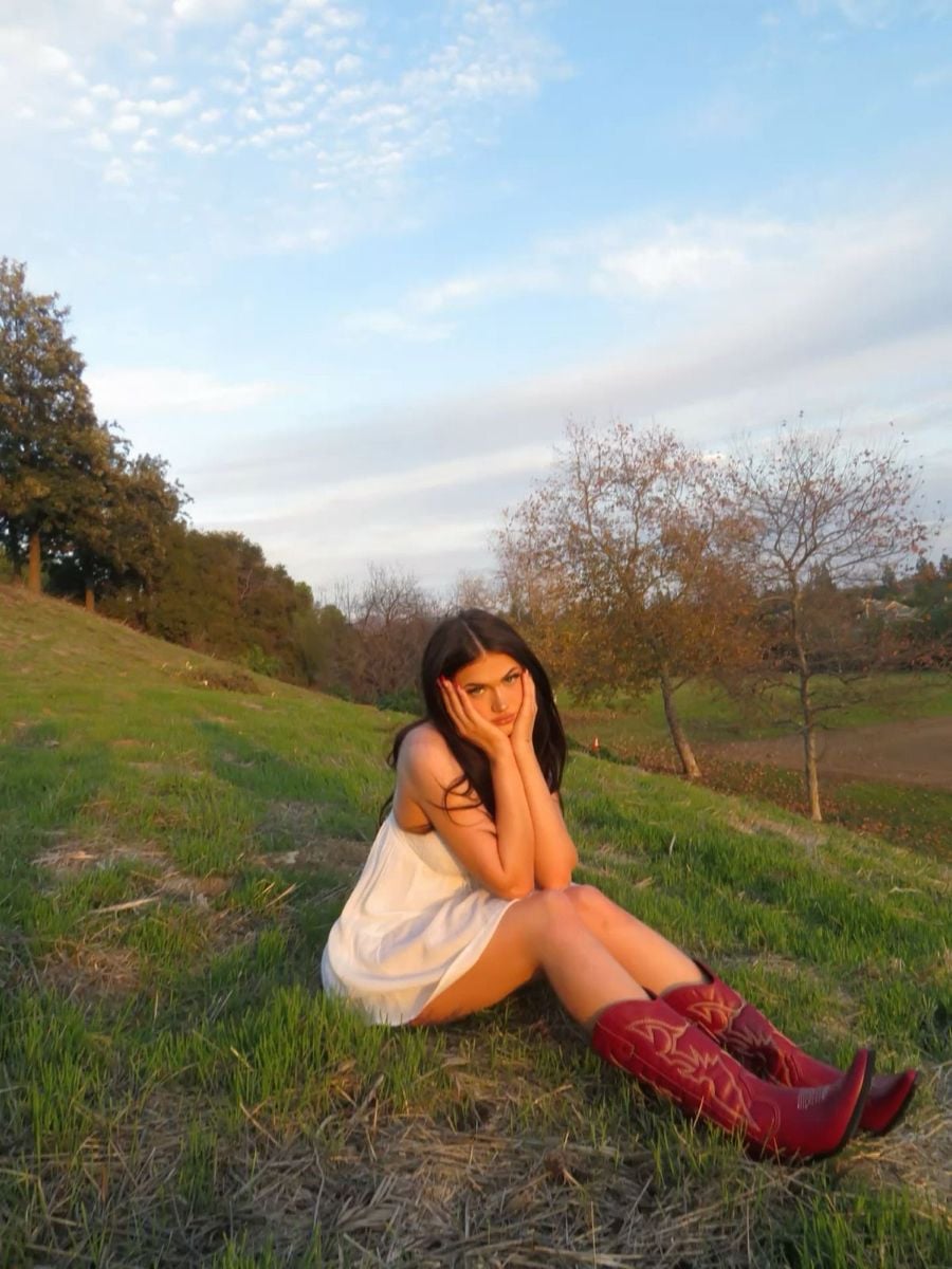 A woman in a white dress and red cowboy boots sits on green grass on a hillside, resting her chin on her hands, with trees and a partly cloudy sky in the background.