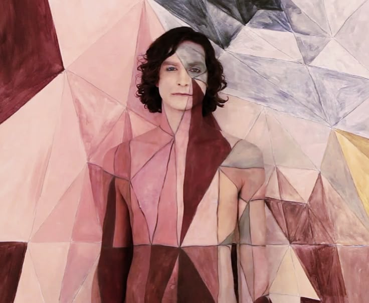 A person with curly hair stands against a wall painted with geometric shapes in muted colors. The person's body is painted to blend seamlessly with the patterned background.
