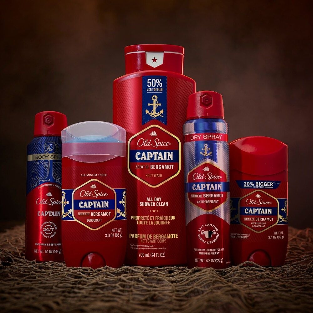 Five Old Spice Captain Scent of Bergamot products, including deodorants, body spray, and shower gel, are arranged upright on a dark textured surface with a brown background.