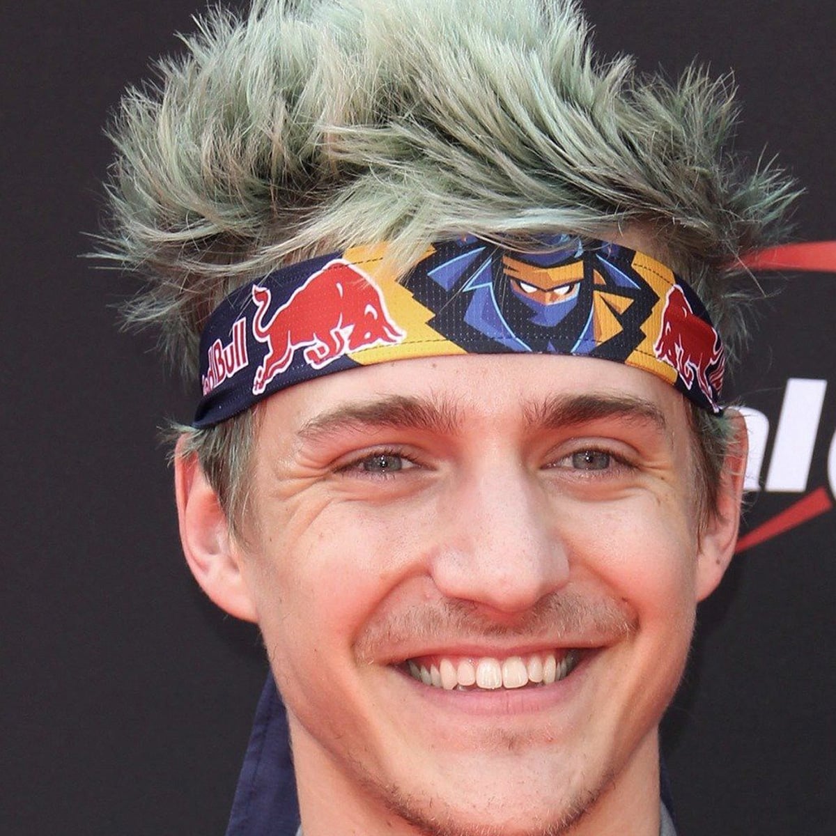 A smiling person with spiky, light blue hair wears a colorful Red Bull headband and stands in front of a dark background with partial white and red text.