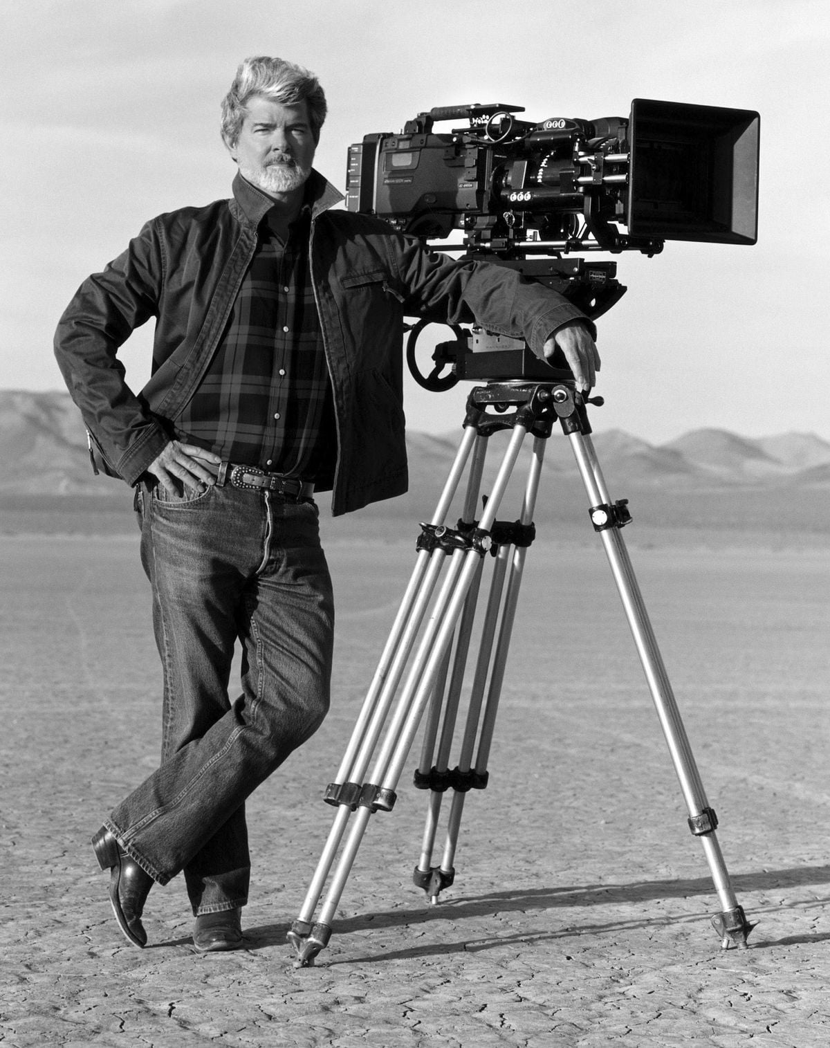 A man with a beard stands outdoors on a dry, cracked landscape, leaning against a large film camera mounted on a tripod, with mountains visible in the background. The image is in black and white.