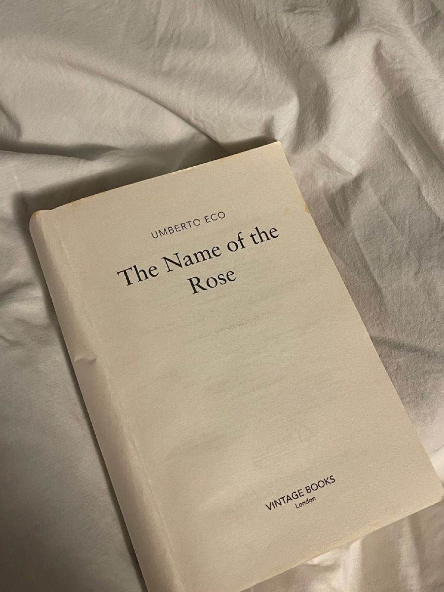 An open book titled "The Name of the Rose" by Umberto Eco lies on a white, wrinkled bedsheet. The publisher, Vintage Books London, is printed at the bottom of the page.