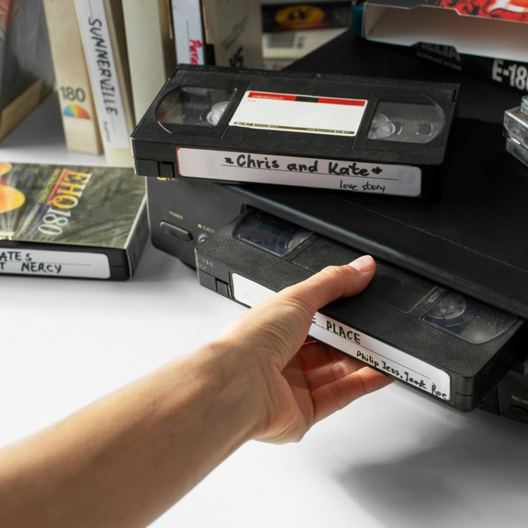A hand is pulling a VHS tape from a stack. Several VHS tapes with handwritten labels, including "Chris and Kate" and "Philip Jess-Jack," are visible on a white surface. Other tapes and boxes are in the background.