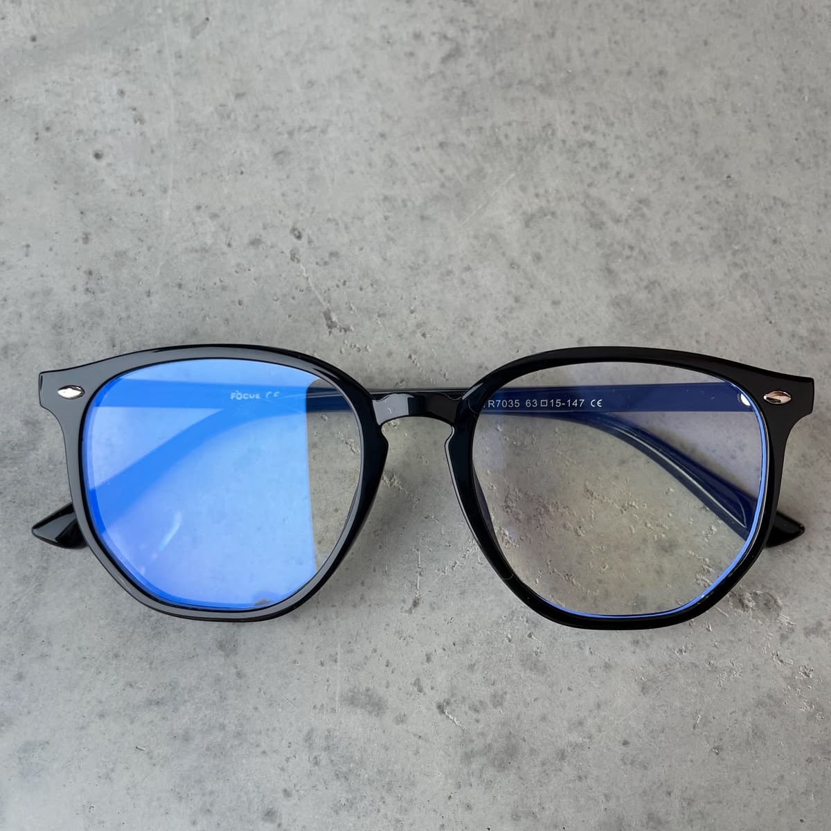 A pair of black-rimmed eyeglasses with blue light reflecting on the lenses, resting on a gray, slightly textured surface.