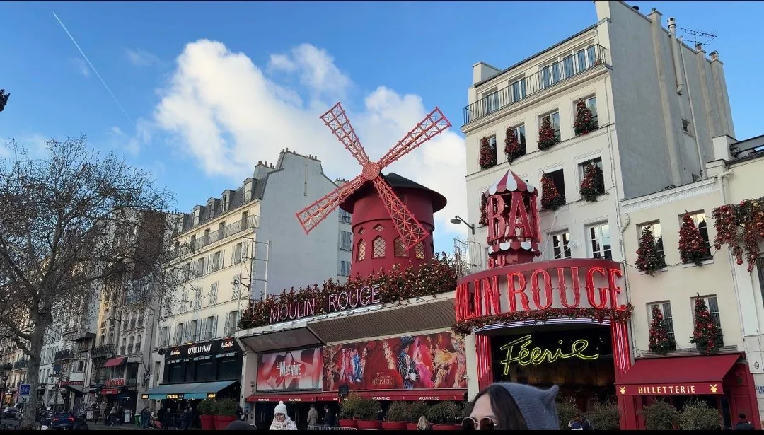The Moulin Rouge cabaret in Paris, featuring its iconic red windmill, vibrant signage, and surrounding buildings, with a blue sky and scattered clouds overhead. People are walking nearby.