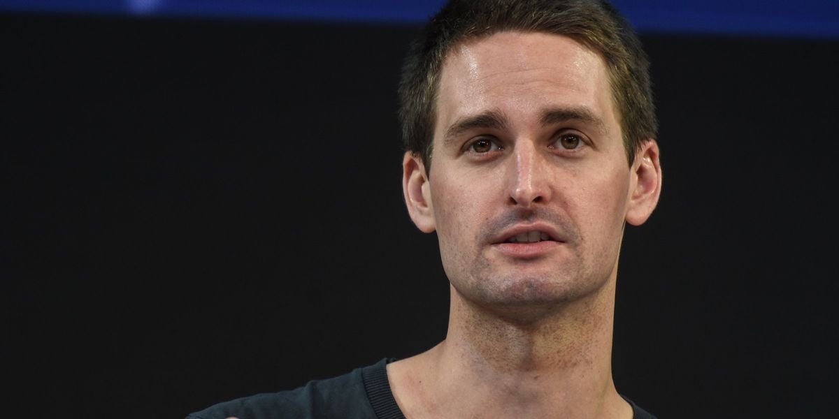 A man with short dark hair is speaking or listening, wearing a dark shirt, in front of a dark background.