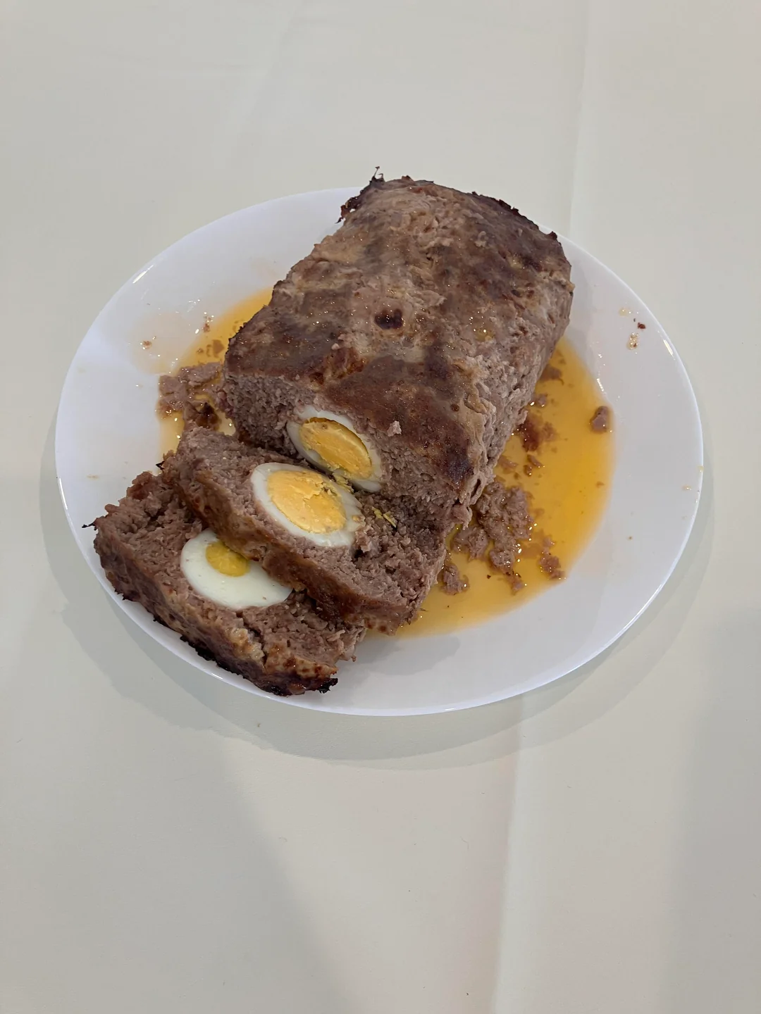 A meatloaf on a white plate with two slices cut, revealing hard-boiled eggs inside. Some orange-brown juices are pooled around the meatloaf on the plate.