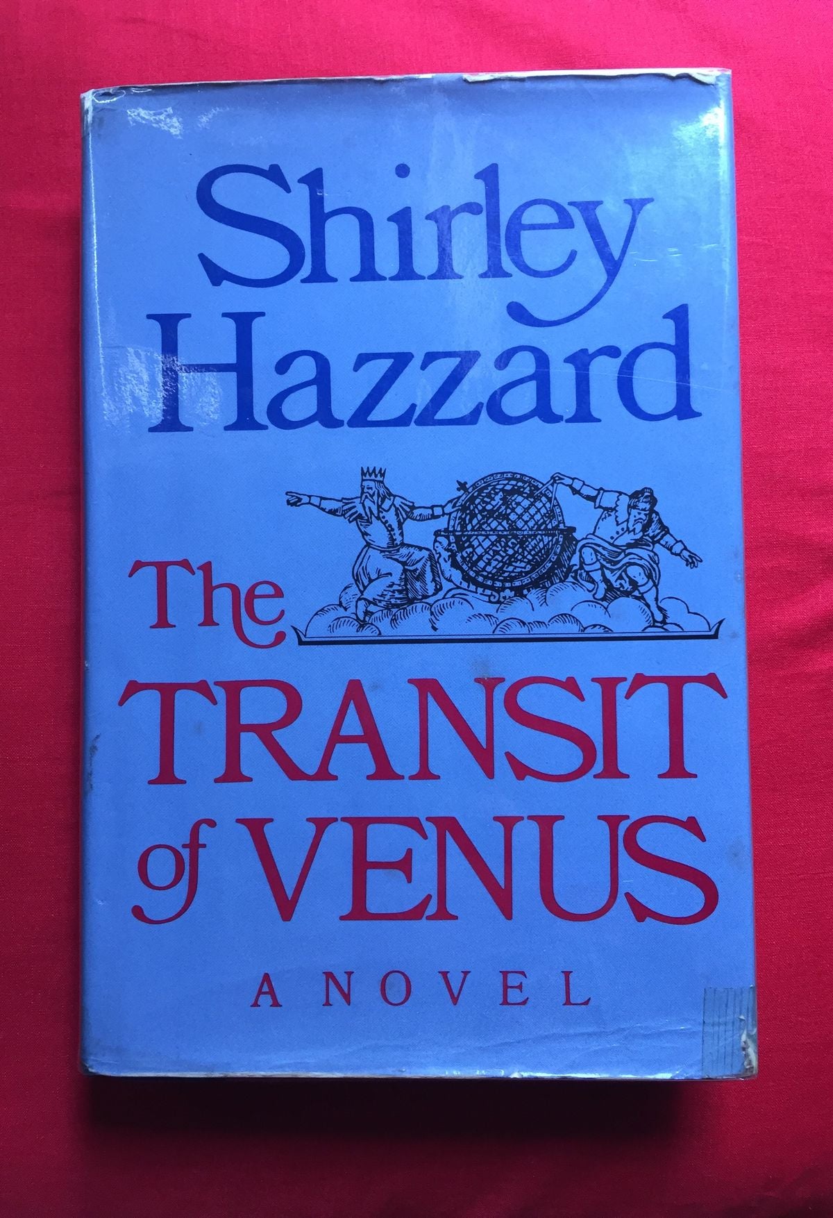 A blue book cover of "The Transit of Venus: A Novel" by Shirley Hazzard, featuring an illustration of two figures pointing at a globe, with the title in large red and blue text.