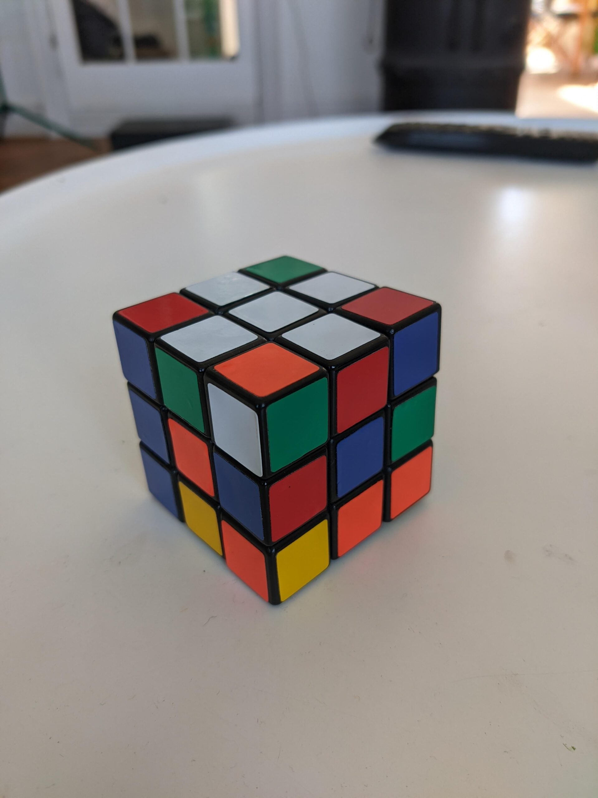 A partially solved Rubik’s Cube with mixed colored squares sits on a round white table, with a remote control and blurred background visible.
