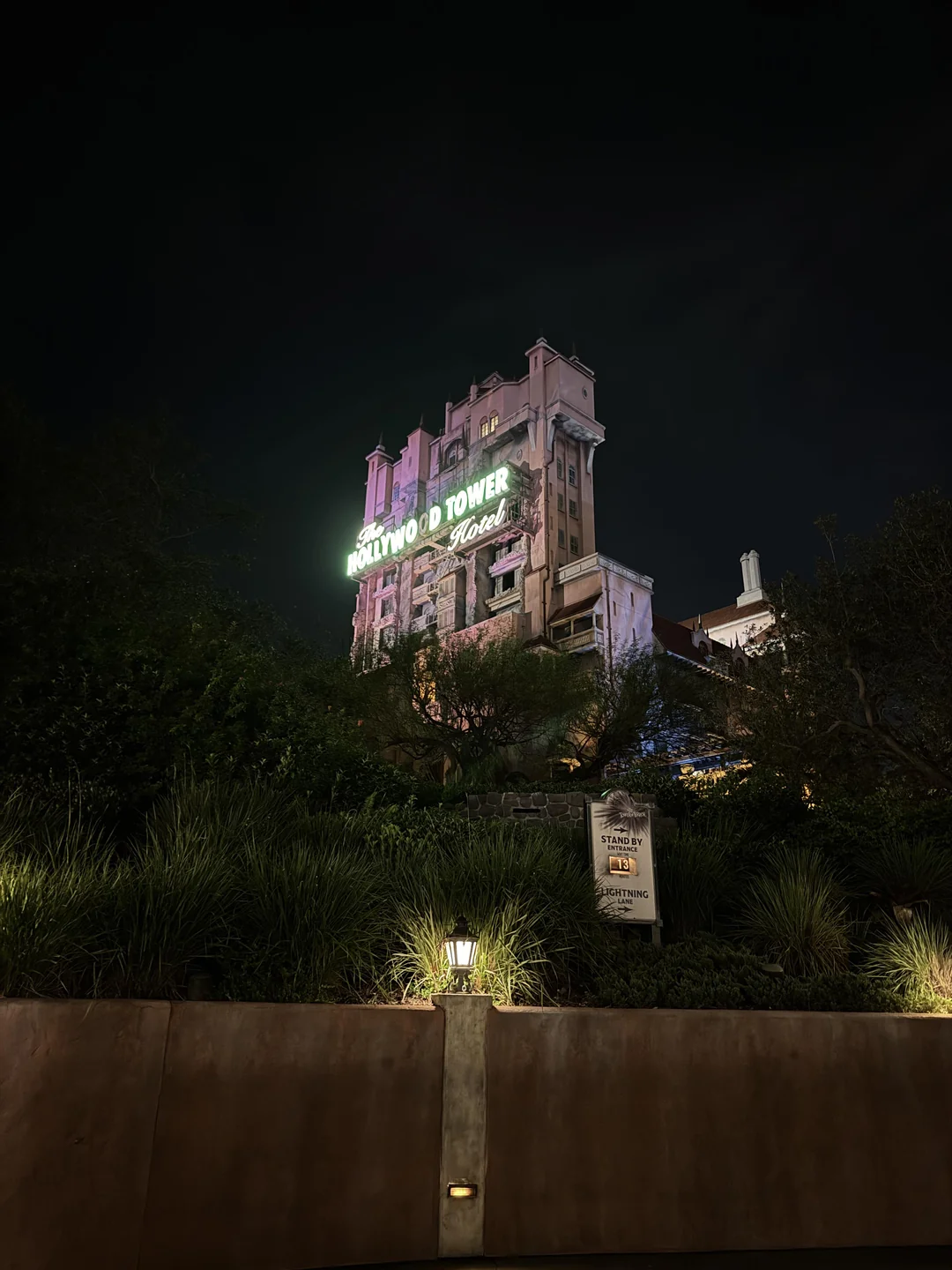 The Hollywood Tower Hotel, illuminated at night with a neon sign, rises above greenery and a lit pathway in front, creating a mysterious and dramatic atmosphere.