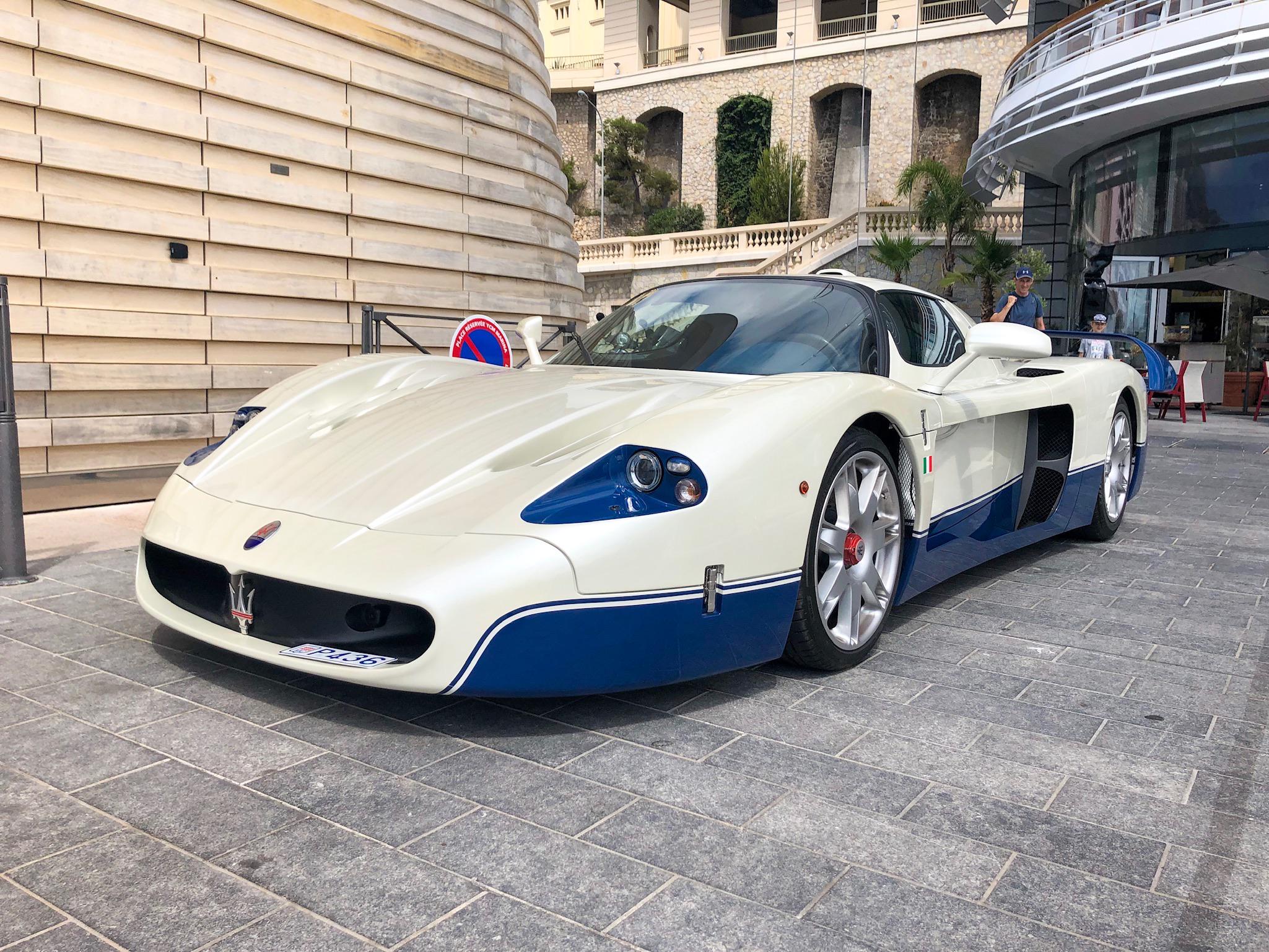 A white and blue Maserati MC12 sports car is parked on a paved street near modern and historic buildings, with a few people visible in the background.